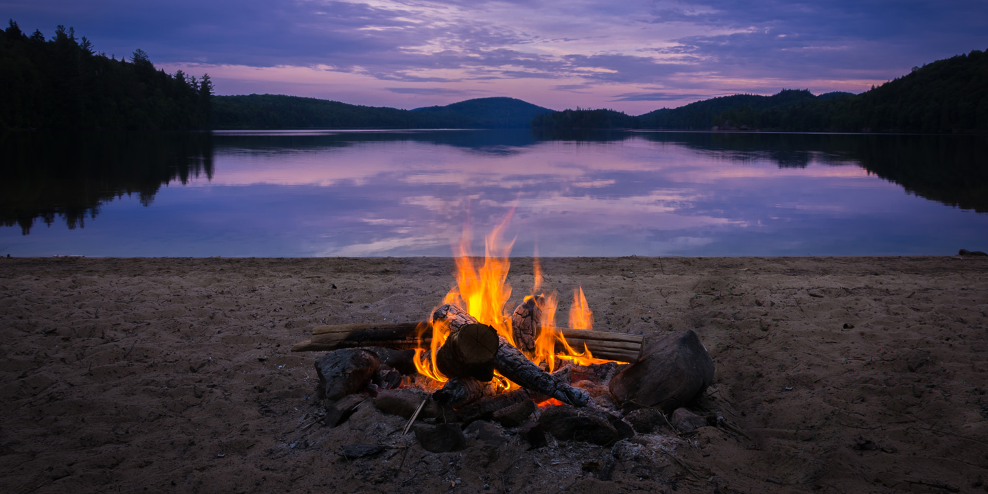 Campfire at the edge of a tranquil lake at sunset.